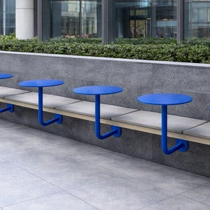 Könnte beinhalten: Außenbereich mit einer langen Bank und fünf kleinen, runden, blauen Tischen. Die Tische werden von gebogenen blauen Metallbeinen getragen. Die Bank hat hellfarbene Kissen und steht vor einer grauen Steinmauer. Grüne Büsche sind im Hintergrund.