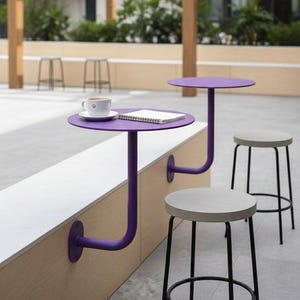 Op de afbeelding: Buitenruimte met paarse, ronde tafels bevestigd aan een witte en houten bank. Een kopje koffie en een notitieboek liggen op een tafel. Grijze krukken met zwarte poten zijn ook aanwezig.