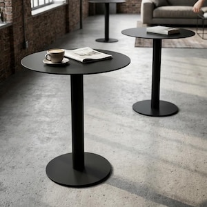 May include: A set of three matte black circular tables, each featuring a single central pedestal base and a flat round top. The foreground table holds a ceramic coffee cup on a saucer and an open newspaper, while another table holds a book.