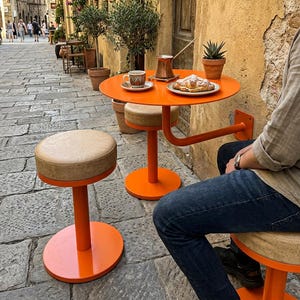 以下が含まれることがあります： オレンジ色のテーブルとスツールがある屋外カフェのシーン。小さな丸いテーブルには、コーヒーカップ、ペストリー、小さな鉢植えの植物が置かれています。カフェは「Cafe Sol」と書かれた看板のある石畳の通りにあります。