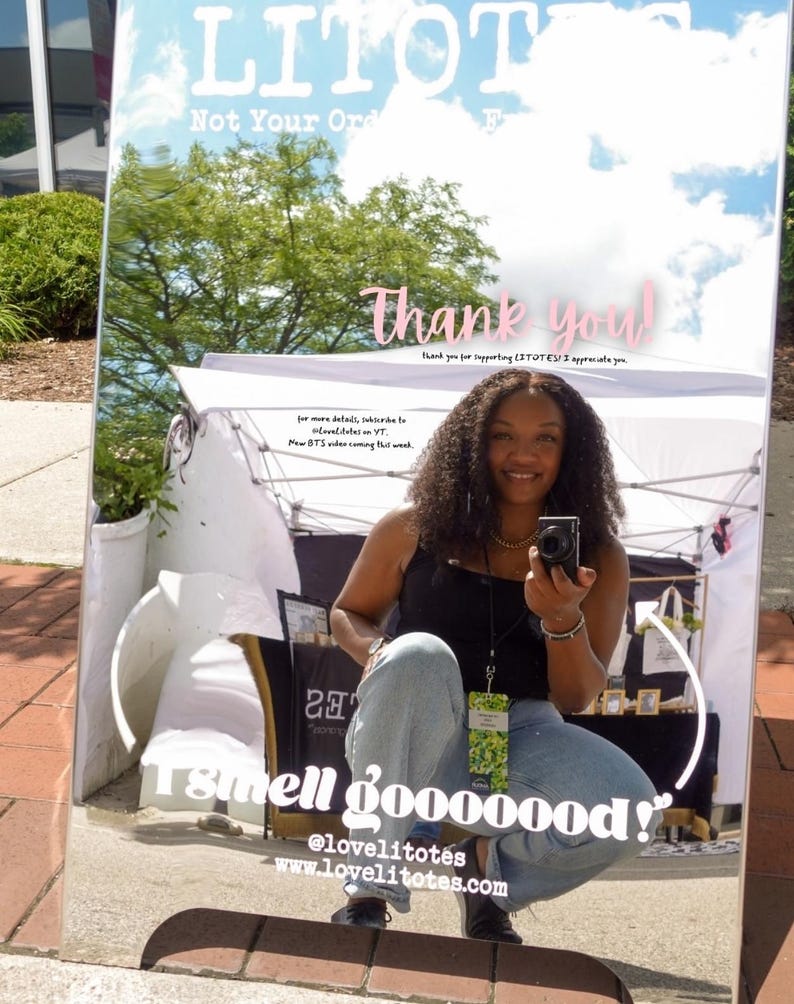 Op de afbeelding: Een groot spiegelbord met de tekst "LITOTES" en "Not Your Ord". Het bord zegt ook "Thank you!" en "I smell gooooood!" Een persoon wordt weerspiegeld in de spiegel, houdt een camera vast en glimlacht.