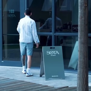 Op de afbeelding: Een buitensc&egrave;ne met een donkergrijs sandwichbord met de tekst "SOTA STUDIO" in het wit. Een persoon loopt naar een glazen deur met het nummer "103". Het bord staat op een bakstenen en houten oppervlak.
