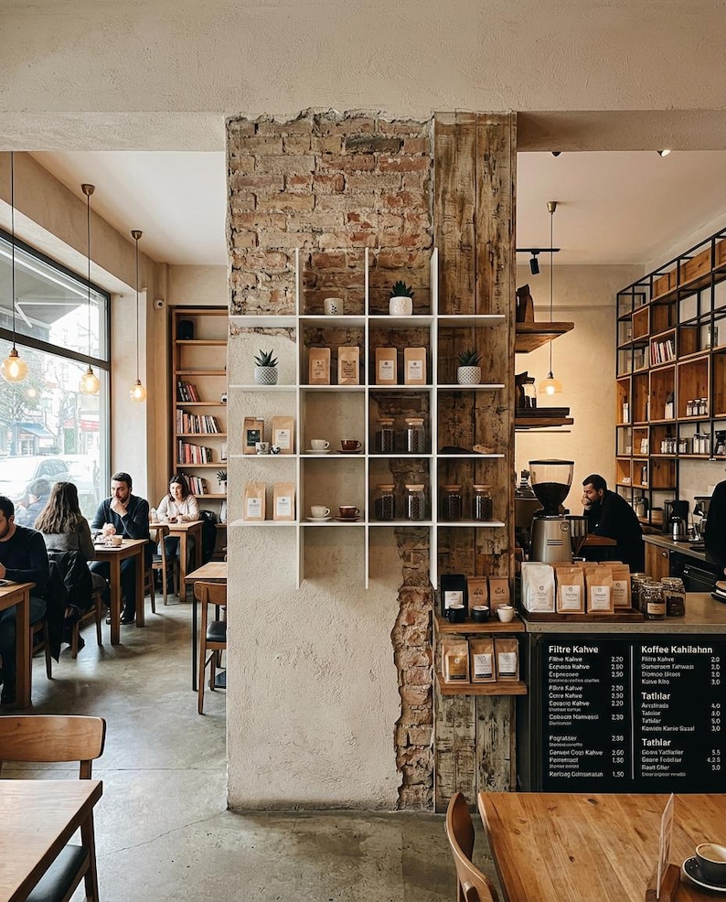 May include: A coffee shop interior featuring a central exposed brick pillar with a white grid shelving unit displaying coffee bags, small potted plants, and ceramic cups. A menu board lists items like Filtre Kahve and Kaffee Kahilahon.