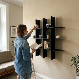 May include: A black wall-mounted grid shelving unit featuring multiple square compartments. The shelves hold various items including vintage cameras, a small potted succulent, a glass candle, and several books arranged in an organized display.