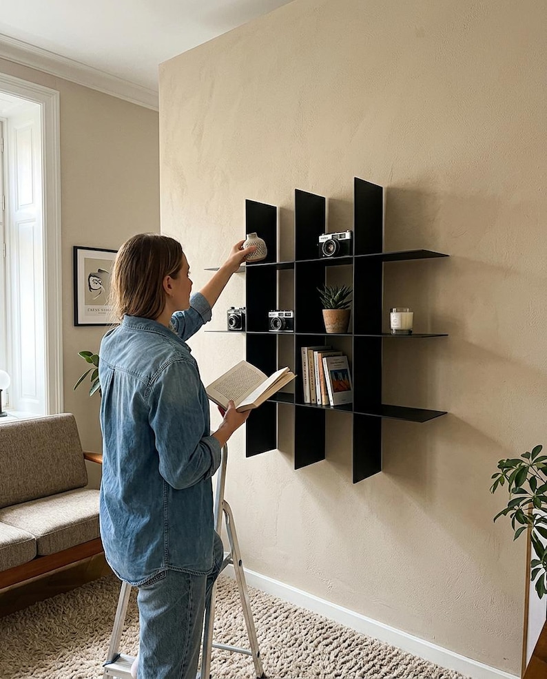 May include: A black wall-mounted grid shelving unit featuring multiple square compartments. The shelves hold various items including vintage cameras, a small potted succulent, a glass candle, and several books arranged in an organized display.