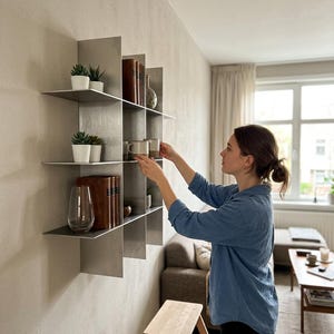 May include: A modular wall-mounted shelving unit made of brushed silver metal with a grid-like structure. The shelves hold small potted green plants, several hardcover books with brown spines, a clear glass vase, and two ceramic mugs.