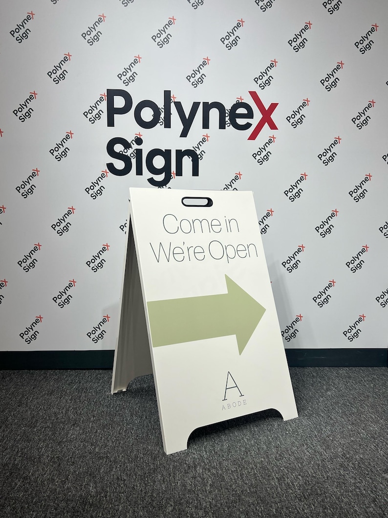 Op de afbeelding: Wit A-frame bord met de tekst "Come in We're Open" en een groene pijl. Het bord heeft een zwarte handgreep en de letter "A" met "ABODE" eronder. De achtergrond toont het "Polynex Sign" logo.