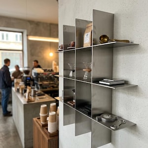 May include: A wall-mounted metal grid shelving unit with a brushed finish, featuring multiple square compartments. The shelves hold various coffee accessories, including a brown paper bag, a brass scoop, glass carafes, a scale, and metal discs.