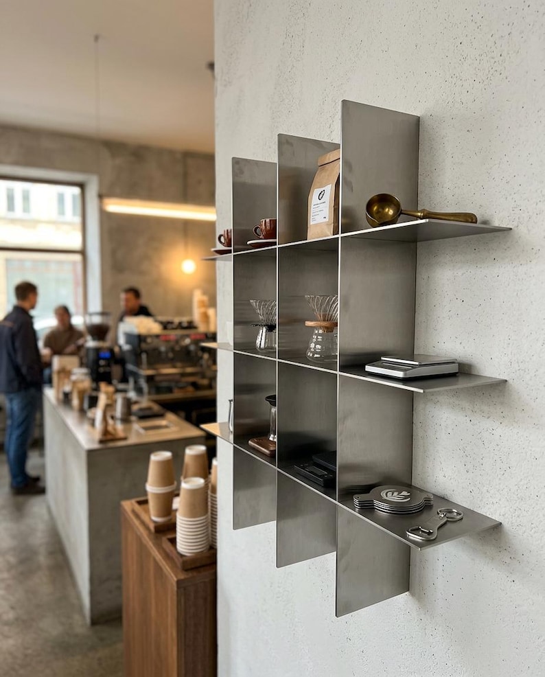 May include: A wall-mounted metal grid shelving unit with a brushed finish, featuring multiple square compartments. The shelves hold various coffee accessories, including a brown paper bag, a brass scoop, glass carafes, a scale, and metal discs.