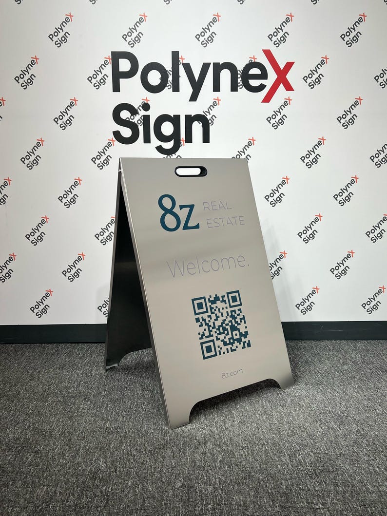 Op de afbeelding: Een zilverkleurig A-framebord met de tekst "8z REAL ESTATE Welcome." en een QR-code. Het bord heeft een handvat aan de bovenkant en wordt weergegeven op een grijze vloerbedekking. De achtergrond toont het "Polynex Sign" logo.