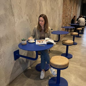 Può includere: Scena di un bar con tavoli rotondi blu e sgabelli abbinati con sedute beige. Una persona è seduta a un tavolo, legge una rivista e beve caffè. I tavoli sono fissati al muro e hanno un design curvo unico.
