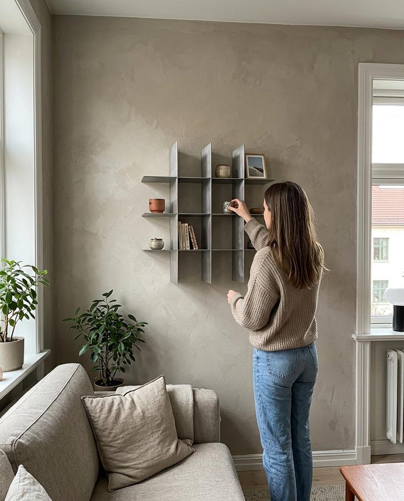 May include: A wall-mounted metal grid shelving unit featuring a series of square compartments. The shelf is constructed from thin, grey vertical and horizontal panels that intersect to create multiple open storage spaces for small decorative items.