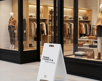 Puede incluir: Un letrero de acera en forma de A de color blanco con un asa recortada en la parte superior. El letrero presenta texto negro que dice "THANKS, I FOUND IT IN Designer Exchange" y un logotipo "DE" en la parte inferior, frente a una tienda.