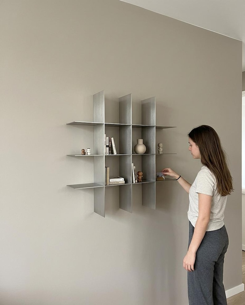 May include: A wall-mounted metal shelving unit featuring a grid design with four vertical dividers and three horizontal shelves, creating twelve open compartments. The shelves are finished in a brushed silver tone and hold various small decorative items.