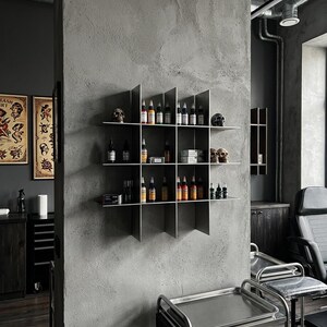 May include: A wall-mounted metal grid shelf featuring four horizontal tiers and four vertical dividers, creating sixteen square compartments. The shelves hold various small bottles of colorful tattoo ink, small boxes, and decorative skull figurines.