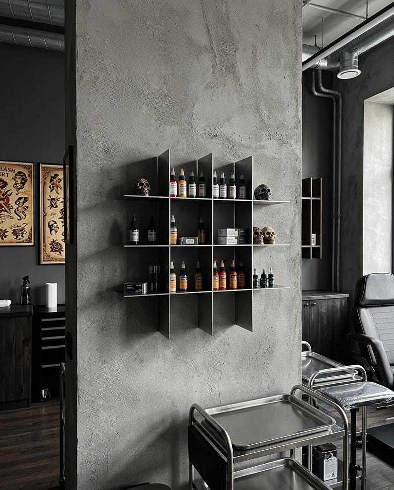 May include: A wall-mounted metal grid shelf featuring four horizontal tiers and four vertical dividers, creating sixteen square compartments. The shelves hold various small bottles of colorful tattoo ink, small boxes, and decorative skull figurines.