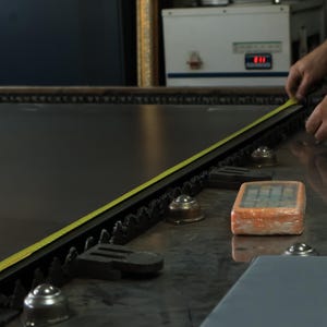 May include: A large, dark, flat industrial sheet of material rests on a metal table with ball bearings. A yellow measuring tape is extended along the edge of the sheet. An orange rectangular control device sits nearby on the metallic surface.
