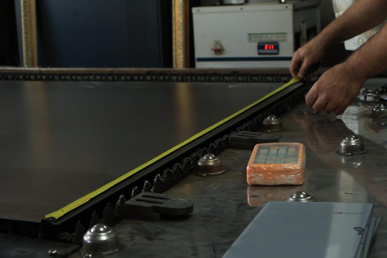 May include: A large, dark, flat industrial sheet of material rests on a metal table with ball bearings. A yellow measuring tape is extended along the edge of the sheet. An orange rectangular control device sits nearby on the metallic surface.