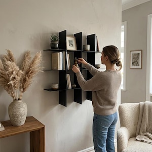 May include: A black metal wall-mounted shelving unit featuring a grid-like design with multiple square compartments. The shelf is mounted on a light-colored wall and holds various books, small decorative objects, and a framed picture.