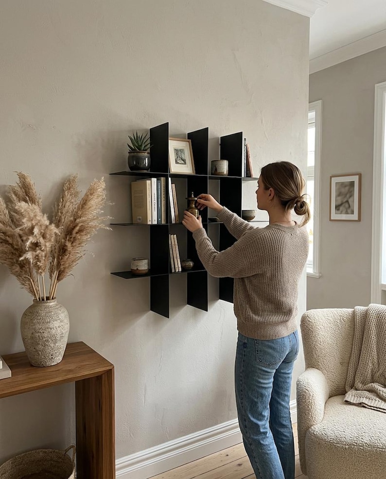 May include: A black metal wall-mounted shelving unit featuring a grid-like design with multiple square compartments. The shelf is mounted on a light-colored wall and holds various books, small decorative objects, and a framed picture.