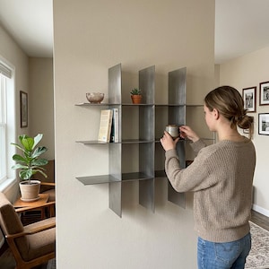 May include: A wall-mounted metal shelving unit featuring a grid design with multiple open compartments. The shelf is constructed from thin, dark gray steel plates arranged in a cross-hatch pattern, providing several flat surfaces for storage and display.