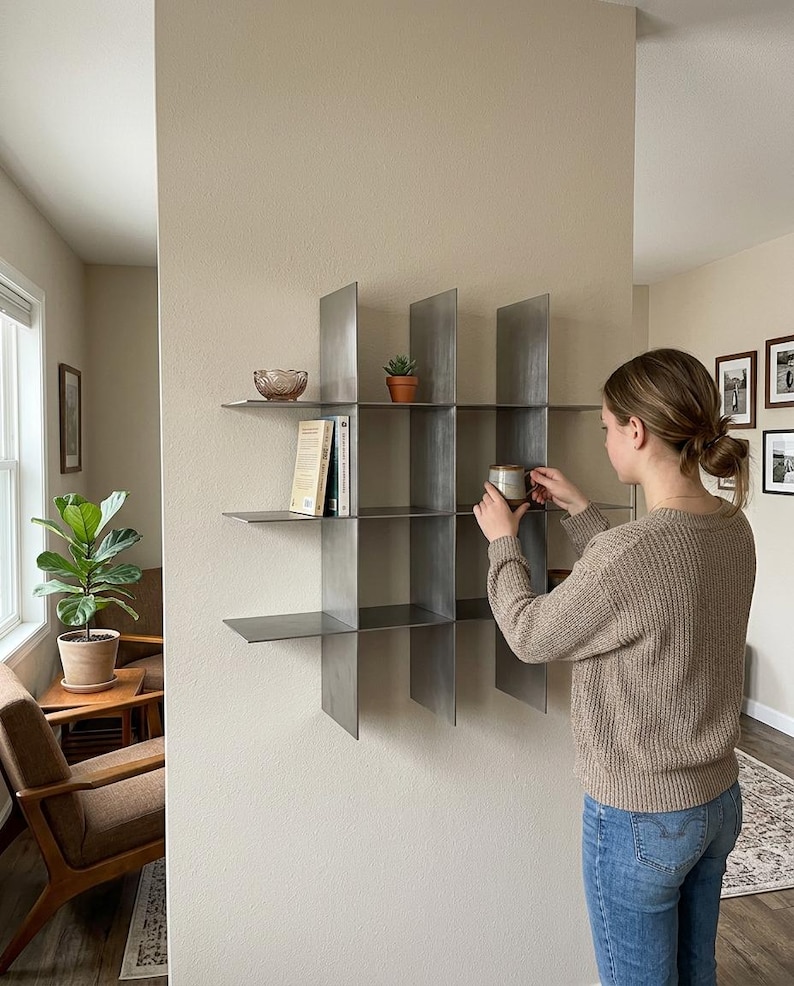 May include: A wall-mounted metal shelving unit featuring a grid design with multiple open compartments. The shelf is constructed from thin, dark gray steel plates arranged in a cross-hatch pattern, providing several flat surfaces for storage and display.