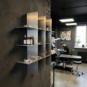 May include: A wall-mounted metal grid shelf with a brushed silver finish, featuring multiple square compartments. The shelves hold various small bottles with black caps, clear circular containers, and metal tattoo equipment components.