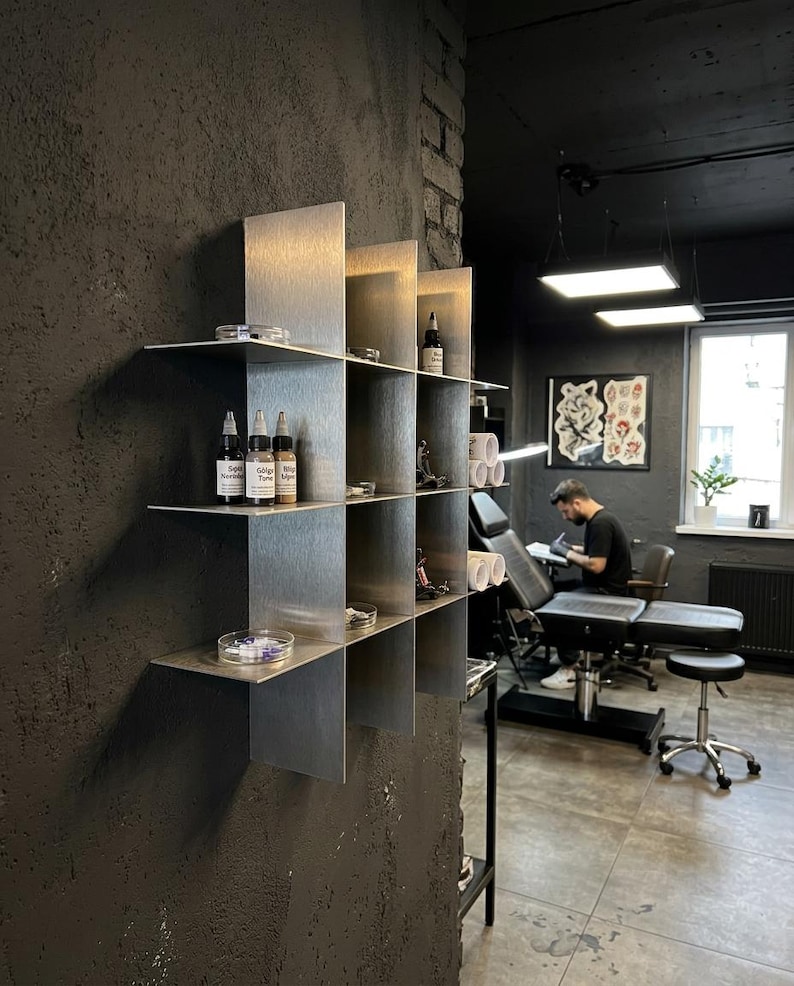 May include: A wall-mounted metal grid shelf with a brushed silver finish, featuring multiple square compartments. The shelves hold various small bottles with black caps, clear circular containers, and metal tattoo equipment components.