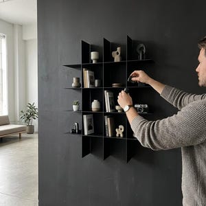 May include: A black metal wall-mounted grid shelving unit featuring sixteen square compartments. The shelves are filled with various decorative objects, including small ceramic vases, books, a vintage camera, and abstract sculptures arranged on the shelves.