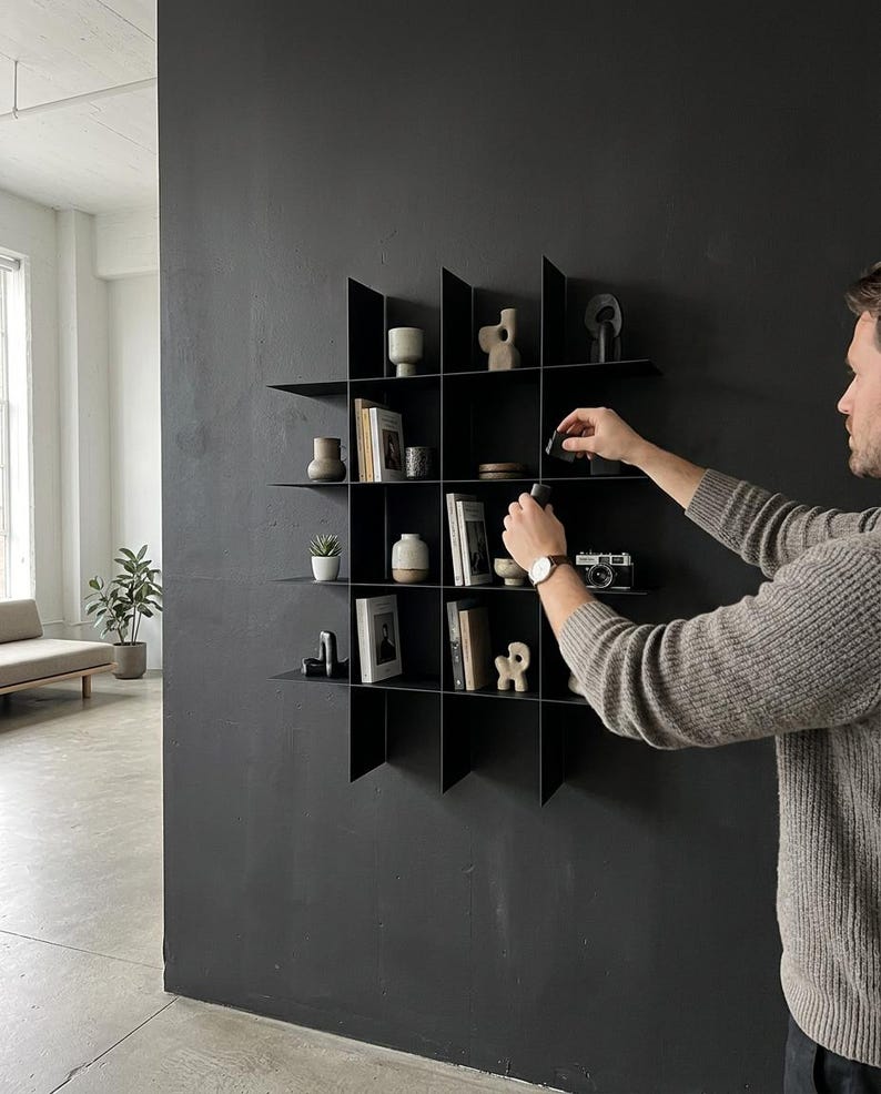 May include: A black metal wall-mounted grid shelving unit featuring sixteen square compartments. The shelves are filled with various decorative objects, including small ceramic vases, books, a vintage camera, and abstract sculptures arranged on the shelves.
