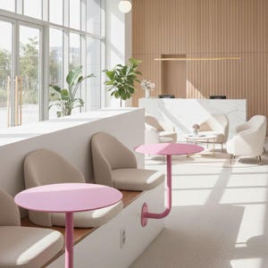 Op de afbeelding: Een modern interieur met een wachtruimte met lichtroze, ronde bijzettafels bevestigd aan een witte bank. Beige stoelen zijn langs de bank geplaatst. De kamer heeft grote ramen, planten en een receptie.