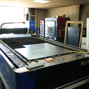 May include: A large industrial laser cutting machine with a blue and white frame, featuring a flat metal bed holding a sheet of steel. The machine includes a control monitor and a gantry system. A yellow measuring tape is held over the metal sheet.
