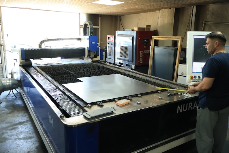 May include: A large industrial laser cutting machine with a blue and white frame, featuring a flat metal bed holding a sheet of steel. The machine includes a control monitor and a gantry system. A yellow measuring tape is held over the metal sheet.