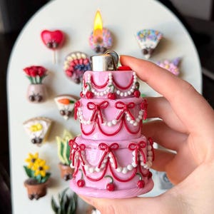 Peut inclure: Un briquet rose en forme de gâteau à trois niveaux, avec des nœuds rouges, des cerises et des perles. Le briquet a une flamme allumée en haut. D'autres aimants décoratifs sont en arrière-plan, dont des fleurs et des cœurs.