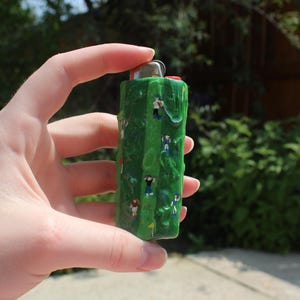 May include: A green lighter with a textured surface, decorated with miniature figures of golfers. The figures are dressed in various colors and are positioned as if playing golf. The lighter has a silver metal top and a red button.
