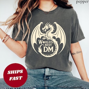 Puede incluir: Camiseta gris oscuro con un gráfico de dragón color crema. El dragón está centrado, con el texto "WORLD'S BEST DM" en un diseño circular. La camiseta es de estilo casual y cómodo.