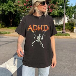 Könnte beinhalten: Schwarzes T-Shirt mit dem Wort "ADHD" in einer feurigen Schriftart. Darunter befindet sich eine Skelettgrafik, die zwei Waffen hält. Das Shirt ist ein lässiges Kurzarm-Design.