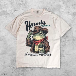 May include: Ivory t-shirt with a cartoon frog dressed as a cowboy, wearing a brown hat, jacket, and red bandana. The frog holds a small potted cactus. Text on the shirt reads "Howdy Pardner... I mean, Partner" in a stylized font.