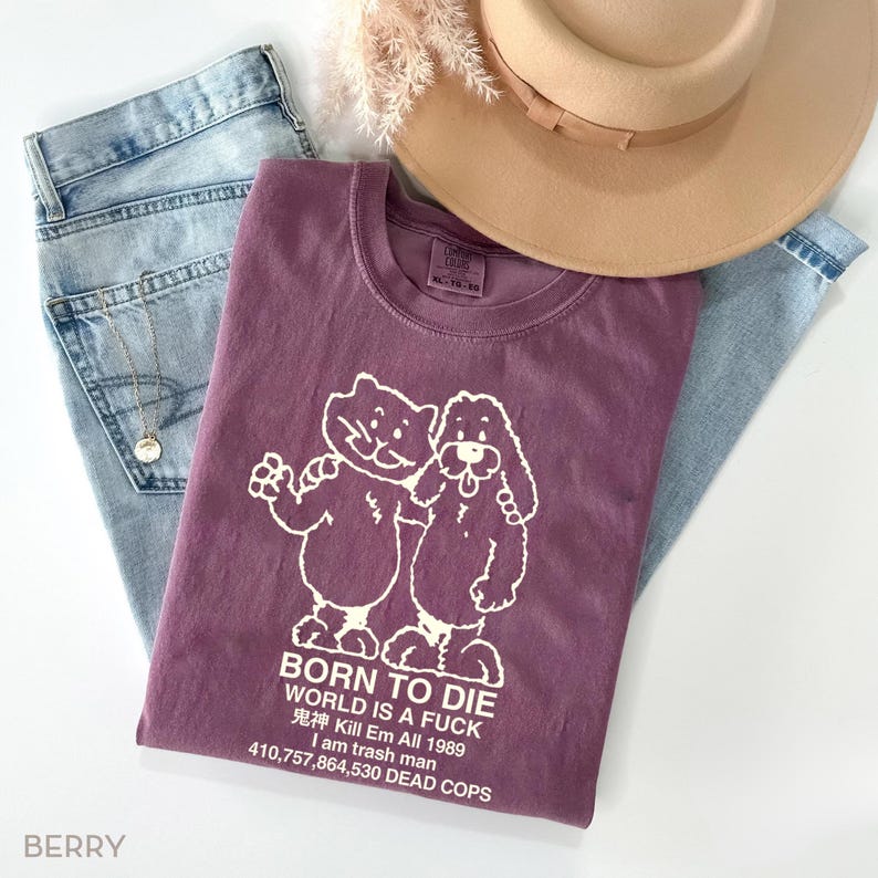 Pode incluir: Uma camiseta cor de baga com um gr&aacute;fico de um gato e um cachorro, e o texto "BORN TO DIE WORLD IS A FUCK 鬼神 Kill Em All 1989 I am trash man 410,757,864,530 DEAD COPS." A camiseta &eacute; combinada com jeans claros e um chap&eacute;u bege.