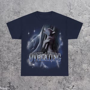 May include: Navy blue t-shirt featuring a graphic of a howling wolf with a mountain backdrop and lightning bolts. The word "OVULATING" is displayed in a stylized font across the bottom of the design.