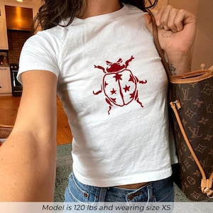 May include: White crop top with a red bug graphic. The bug design features a detailed outline with star accents. The shirt is paired with blue jeans. The model is wearing size XS.