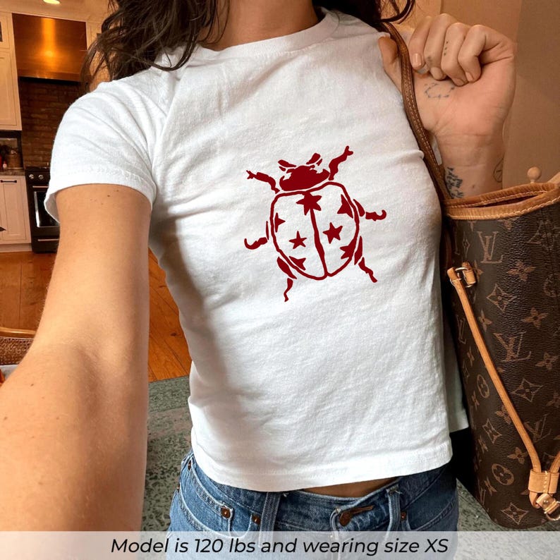 Op de afbeelding: Witte crop top met een rode keverafbeelding. Het keverontwerp heeft een gedetailleerde omtrek met steraccenten. Het shirt wordt gecombineerd met een blauwe jeans. Het model draagt maat XS.