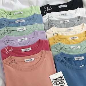 Pode incluir: Uma cole&ccedil;&atilde;o de camisetas dobradas em v&aacute;rias cores, incluindo preto, branco e cinza. Cada camisa tem gola redonda e uma pequena etiqueta. As cores s&atilde;o rotuladas com escrita &agrave; m&atilde;o, e uma etiqueta diz "COMFORT COLORS."