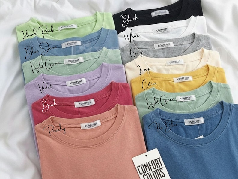 Pode incluir: Uma cole&ccedil;&atilde;o de camisetas dobradas em v&aacute;rias cores, incluindo preto, branco e cinza. Cada camisa tem gola redonda e uma pequena etiqueta. As cores s&atilde;o rotuladas com escrita &agrave; m&atilde;o, e uma etiqueta diz "COMFORT COLORS."