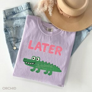 Pu&ograve; includere: Una maglietta lilla con la scritta "LATER" in rosa e un alligatore verde a cartoni animati. La maglietta fa parte di un abbigliamento casual, con jeans azzurri e un cappello beige.