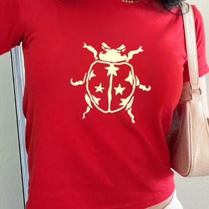Op de afbeelding: Een rood T-shirt met een cr&egrave;mekleurige keverafbeelding met sterdetails. De top met korte mouwen is gemaakt van zacht materiaal en is geschikt voor casual wear.