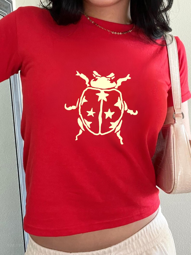 Op de afbeelding: Een rood T-shirt met een cr&egrave;mekleurige keverafbeelding met sterdetails. De top met korte mouwen is gemaakt van zacht materiaal en is geschikt voor casual wear.