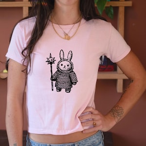 Może przedstawiać: Jasnoróżowy crop top z czarno-białą ilustracją królika w zbroi kolczugowej trzymającego maczugę w kształcie gwiazdy. Królik ma długie uszy i poważny wyraz twarzy. Koszulka ma krótkie rękawy.