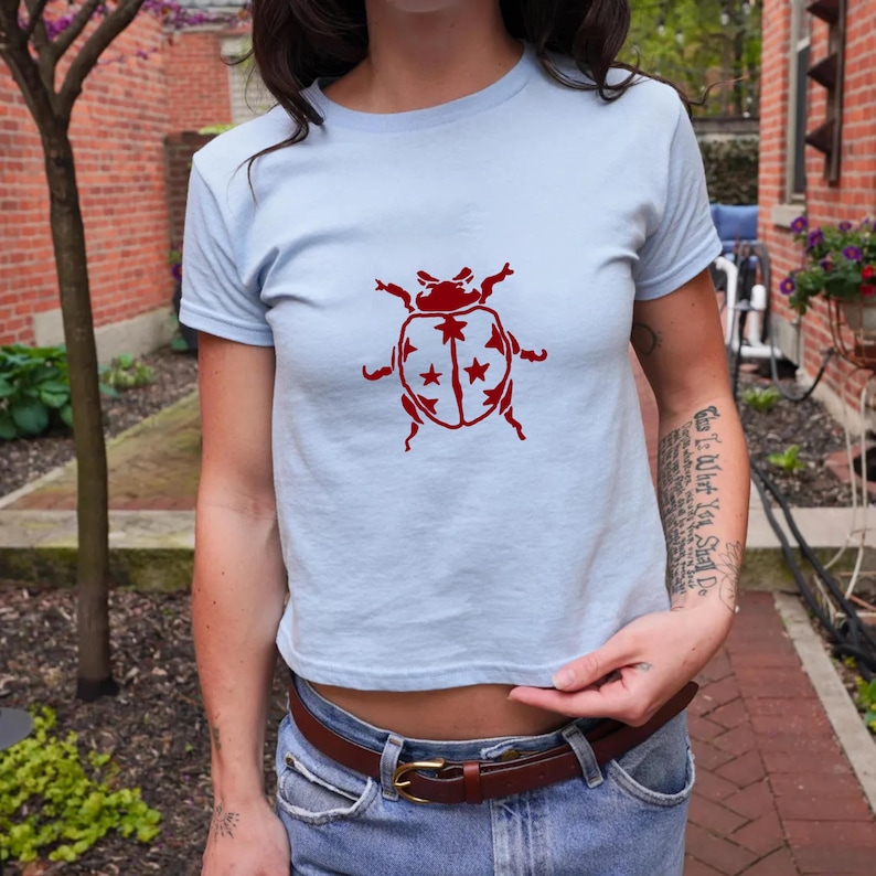 Op de afbeelding: Lichtblauw crop top met een rode insectenafbeelding met sterren. Het T-shirt heeft korte mouwen en een ronde hals. Het insectenontwerp staat in het midden van de voorkant. Het model draagt een blauwe spijkerbroek en een bruine riem.