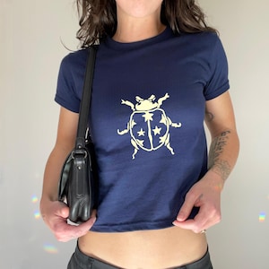 Op de afbeelding: Marineblauwe t-shirt met een cr&egrave;mekleurige lieveheersbeestje-afbeelding met sterren. De top met korte mouwen heeft een ronde hals. Het shirt is gemaakt van zacht materiaal.